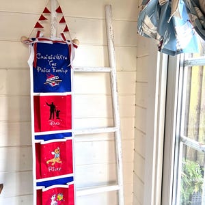 May include: A white wooden ladder leaning against a wall with a red, white, and blue fabric organizer hanging on it. The organizer has four pockets with embroidered characters from Disney movies. The top pocket has the text "Cruisin' With The Price Family". The second pocket has the text "Dad". The third pocket has the text "Riley". The fourth pocket has the text "Avery".