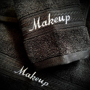 Könnte beinhalten: Nahaufnahme von zwei dunkelgrauen Handtüchern mit dem Wort "Makeup" in weißer Schrift gestickt. Die Handtücher haben eine strukturierte Oberfläche und eine dekorative Bordüre.