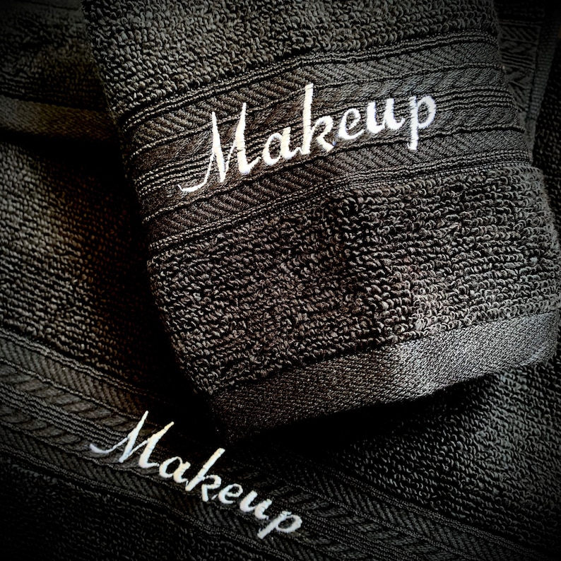 May include: Close-up of two dark grey towels with the word "Makeup" embroidered in white script. The towels have a textured surface and a decorative border.