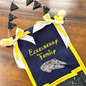 May include: A navy blue fabric wall organizer with yellow trim and a star wars theme. It has a pocket for mail, a pocket for keys, and a pocket for other items. The organizer has a white ribbon bow and a string of black and white triangle flags. The text "Eckelberry Family" is embroidered on the organizer.