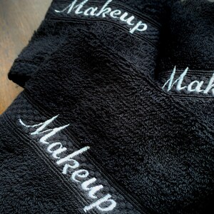 May include: Three black washcloths embroidered with the word "Makeup" in white script. The towels are made of a soft, absorbent material, ideal for removing makeup. The towels are folded and stacked on top of each other.