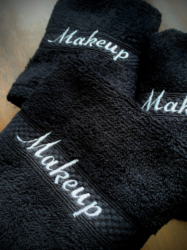 May include: Three black washcloths embroidered with the word "Makeup" in white script. The towels are made of a soft, absorbent material, ideal for removing makeup. The towels are folded and stacked on top of each other.