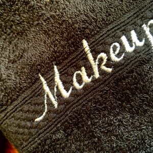 May include: Close-up of a black, plush towel with the word "Makeup" embroidered in white script. The towel has a textured surface and a decorative border. Suitable for makeup removal and skincare routines.