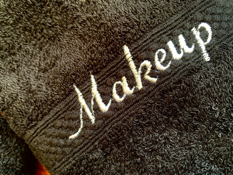 May include: Close-up of a black, plush towel with the word "Makeup" embroidered in white script. The towel has a textured surface and a decorative border. Suitable for makeup removal and skincare routines.