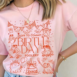 May include: A light pink t-shirt with a red graphic featuring frogs, party hats, and the text "OFF TO COMMIT BIRTHDAY FOOLERY." The design includes a birthday cake and the name "KAREN."