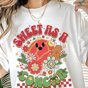 May include: White t-shirt with a retro Christmas graphic. The design features the text "Sweet as a Song" in red and green, along with a red ornament, gingerbread man, flowers, and stars. A festive holiday tee.