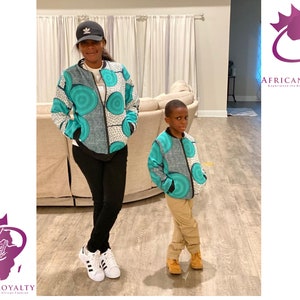 May include: A woman and a child wearing matching teal, white, and black patterned bomber jackets. The woman is wearing a black baseball cap and black pants. The child is wearing khaki pants. The text "AFRICAN ROYALTY" is visible in the image.