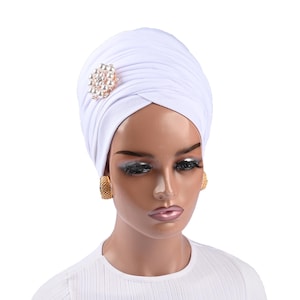 Puede incluir: Turbante blanco con un broche de perlas y oro. El turbante está hecho de una tela suave y elástica y está diseñado para ser usado cómodamente.