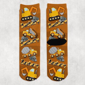 May include: A pair of brown socks featuring construction vehicles like excavators, dump trucks, and cement mixers. The name "Zachary" is printed in yellow. The socks have a construction theme.