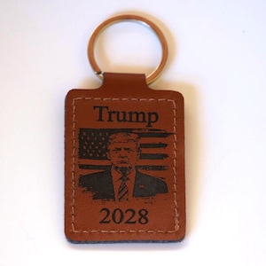 May include: Brown leather keychain with a gold-colored ring. The rectangular tag features the name "Trump", an image of a man, the American flag, and the year "2028" in black.