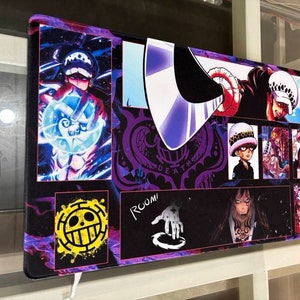 Peut inclure: Un tapis de souris color&eacute; repr&eacute;sentant des personnages de l'anime One Piece. Le tapis de souris a un fond violet, noir et blanc avec des images de diff&eacute;rents personnages, dont un cr&acirc;ne avec un chapeau de paille, un personnage avec une &eacute;p&eacute;e et un personnage avec un cr&acirc;ne et des os crois&eacute;s. Le texte "ROOM!" est visible dans le coin inf&eacute;rieur droit.