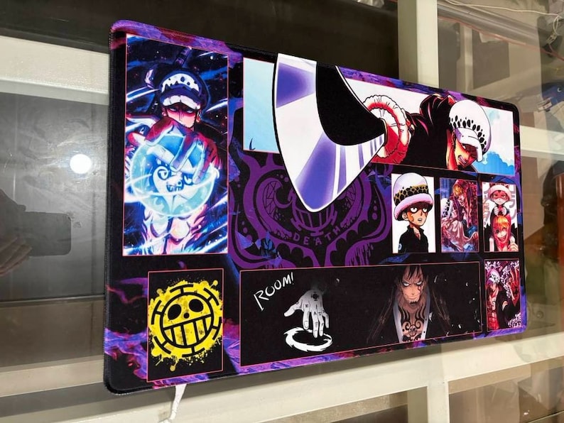 Peut inclure: Un tapis de souris color&eacute; repr&eacute;sentant des personnages de l'anime One Piece. Le tapis de souris a un fond violet, noir et blanc avec des images de diff&eacute;rents personnages, dont un cr&acirc;ne avec un chapeau de paille, un personnage avec une &eacute;p&eacute;e et un personnage avec un cr&acirc;ne et des os crois&eacute;s. Le texte "ROOM!" est visible dans le coin inf&eacute;rieur droit.