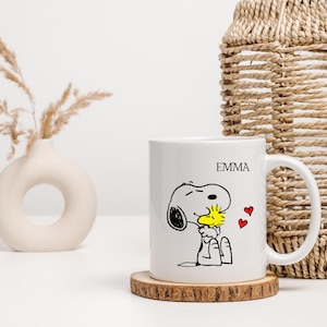 Könnte beinhalten: Weiße Keramiktasse mit einer Schwarzweißillustration von Snoopy, der Woodstock umarmt. Die Tasse trägt den Namen "EMMA" in schwarzer Schrift. Auf der Tasse befinden sich drei rote Herzen.