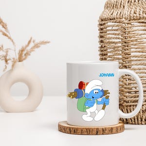 Könnte beinhalten: Weiße Keramiktasse mit einer blauen Comicfigur, die einen Rucksack und einen Stock trägt. Die Figur heißt Schlumpf. Die Tasse trägt den Namen "JOHANN" aufgedruckt.