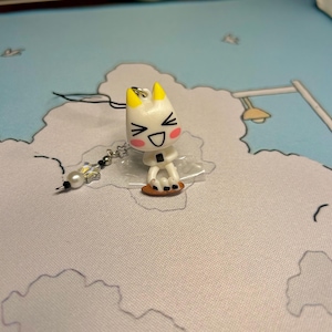 Puede incluir: Encanto de personaje de dibujos animados blanco y amarillo con una cara sonriente y ojos negros. El encanto est&aacute; unido a una cadena plateada con una cuenta de cristal transparente.
