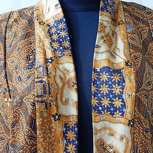Può includere: Un kimono con motivo marrone e blu con un lungo davanti aperto. Il kimono presenta un design batik tradizionale con motivi floreali e geometrici.