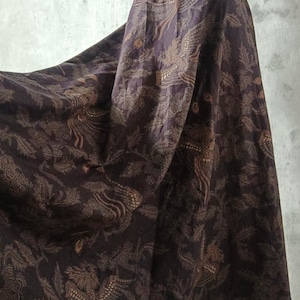 Puede incluir: Una falda larga de color marrón oscuro con un patrón repetido de pájaros y follaje. La tela es ricamente estampada y cae con elegancia. La falda se muestra en un maniquí, mostrando su longitud completa y el intrincado diseño.