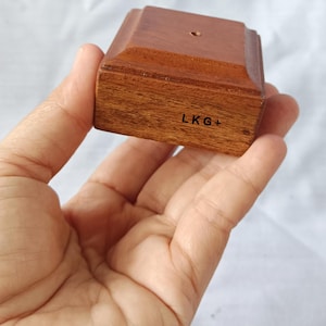 May include: A small, square, wooden display stand with a hole in the top. The stand is made of dark brown wood and has a light brown stain. The bottom of the stand has the text "LKG+" printed on it.