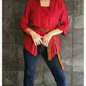 Puede incluir: Una mujer con un top rojo con un cinturón estampado y pantalones vaqueros azules. Está de pie frente a una pared de azulejos.