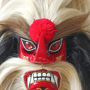 Könnte beinhalten: Eine rote Maske mit glänzendem Finish, weißen Zähnen, schwarz-goldenen Bemalungen und roten Augen. Die Maske ist mit einem leuchtend roten Wollpompon auf der Stirn verziert und von langem, dichtem, cremefarbenem und schwarzem Kunsthaar umgeben.