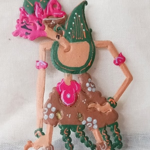 May include: A hand-painted wooden puppet with a green, pink, and brown color scheme. The puppet is a traditional Indonesian shadow puppet, known as a wayang kulit. The puppet is in a standing pose with its arms outstretched. The puppet has a crown and a long, flowing skirt.