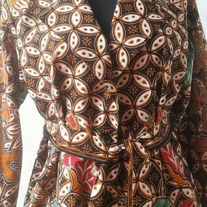 Op de afbeelding: Een bruine en witte gebloemde badjas met lange mouwen en een strikceintuur. De badjas heeft een kraag en zakken.