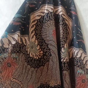 May include: A full-length skirt with a batik design. The skirt features a dark background with intricate patterns in shades of brown, beige, red, and green. The text "LKG+" is visible at the top. The skirt has a wide, flowing shape.