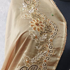 Op de afbeelding: Een beige sjaal met ingewikkelde, geborduurde kanten details. Het kant heeft bloemen- en paisleypatronen met gouden en witte draad, geaccentueerd met kleine kralen. De sjaal is over een zwarte mannequin gedrapeerd.