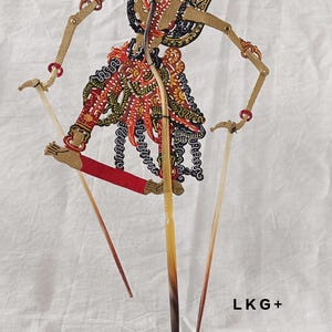 May include: A traditional Indonesian shadow puppet, or wayang kulit, depicting a mythical figure. The puppet is made of intricately carved and painted leather and is mounted on a wooden stand with bamboo rods.