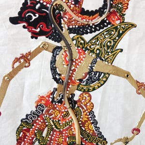 Puede incluir: Un títere de sombras tradicional indonesio, conocido como wayang kulit, que representa a un personaje con detalles intrincados y colores vibrantes. El títere está hecho de cuero y está pintado con detalles dorados, rojos, negros y verdes. El títere está montado en un soporte de madera y está listo para ser utilizado en una representación de títeres de sombras.