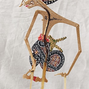 Puede incluir: Un títere de sombras tradicional indonesio, o wayang kulit, que representa a un personaje con un tocado negro y dorado, una pieza de pecho roja y dorada y una falda con estampado en blanco y negro. El títere está hecho de cuero y está montado en un palo de madera.