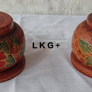 May include: Two wooden containers with intricate floral designs painted in red, green, and brown. The containers have a round shape and a small hole in the top.