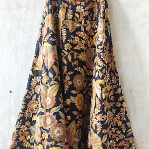 Pode incluir: Uma saia longa e fluida com fundo azul marinho escuro, adornada com um vibrante padrão floral e de pássaros em tons de amarelo, laranja e castanho. O texto "LKG+" é visível no topo.