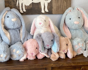 May include: Five plush bunny rabbits in various colors and sizes. The bunnies are arranged in a row on a wooden shelf. The bunnies are all wearing a bow tie. The text "Southern Sass & Shenanigans" is visible on a wooden sign in the background.