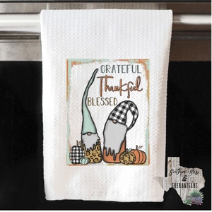May include: A white kitchen towel with a colourful design featuring two gnomes, pumpkins, and the text "Grateful Thankful Blessed".