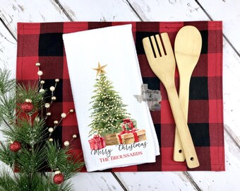 May include: A white Christmas kitchen towel with a watercolor Christmas tree design, gifts, and the text "Merry Christmas". Includes wooden utensils and a red and black buffalo check placemat. Festive holiday decor.