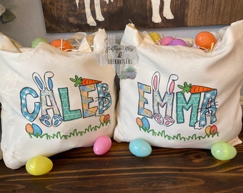 May include: Two white canvas tote bags with personalized Easter bunny designs. The bag on the left says "Caleb" and the bag on the right says "Emma".