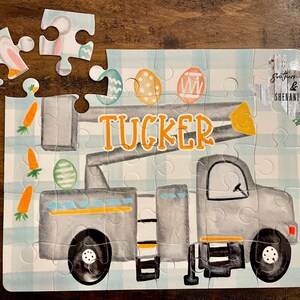 May include: A gray and yellow toy truck with a lift, decorated with Easter eggs and carrots. The truck has the name "TUCKER" written on the side. The puzzle is on a blue and white checkered background.