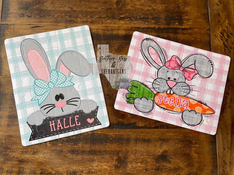 May include: Two personalized Easter bunny jigsaw puzzles. The first puzzle features a gray bunny with a blue bow and the name "Halle" on a black banner. The second puzzle features a gray bunny with a pink bow holding a carrot with the name "Stella" on it.