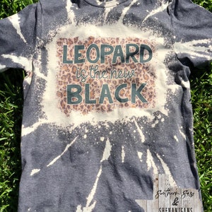 Peut inclure: Un t-shirt gris foncé avec un motif de blanchiment blanc. Le t-shirt porte le texte "LEOPARD is the new BLACK" avec un fond imprimé léopard.