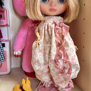 Op de afbeelding: Een pop met blond haar en grote blauwe ogen, gekleed in een jurk met bloemenprint en roze accenten. Een houten trekdier in de vorm van een paard staat op de voorgrond. Een roze teddybeer staat op de achtergrond.