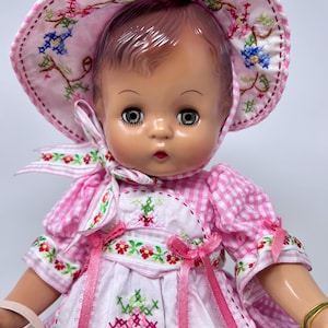 May include: A vinyl doll with brown hair and grey eyes wearing a pink and white gingham dress and matching bonnet. The outfit features white fabric panels with colorful floral cross-stitch embroidery and pink ribbon accents. A gold tag is visible.