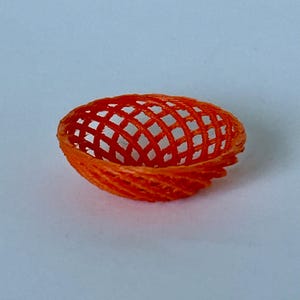 May include: A small, orange, 3D printed bowl with a woven pattern. The bowl is made of plastic and has a round shape.