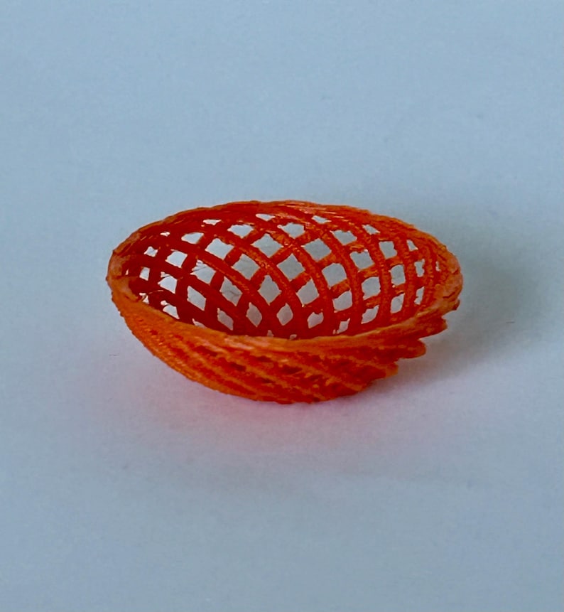 May include: A small, orange, 3D printed bowl with a woven pattern. The bowl is made of plastic and has a round shape.