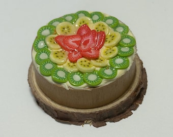 May include: A miniature cake with a fruit topping, including kiwi, banana, and a red strawberry. The cake sits on a wooden base, and the fruit slices are arranged in a decorative pattern. The cake is a light brown color.