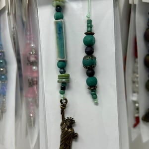 Puede incluir: Dos collares de cuentas verdes y marrones con una pequeña estatua de bronce de la Estatua de la Libertad colgando de uno de los collares. Los collares están empaquetados en bolsas de plástico transparentes con una tarjeta blanca que dice "Primrose & Thyme Doll Boutique".