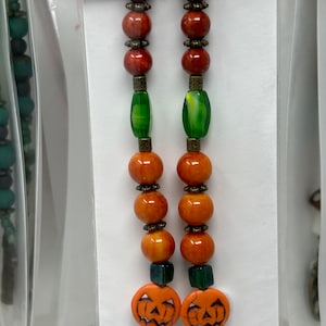 May include: A pair of orange and green beaded earrings with pumpkin-shaped charms. The earrings are made with orange, red, and green beads, and the charms are carved with a jack-o'-lantern face.