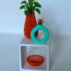 May include: A white cube shelf with three decorative items: a tall orange vase with a green plant, a turquoise vase with a red plant, and a small orange wire basket.