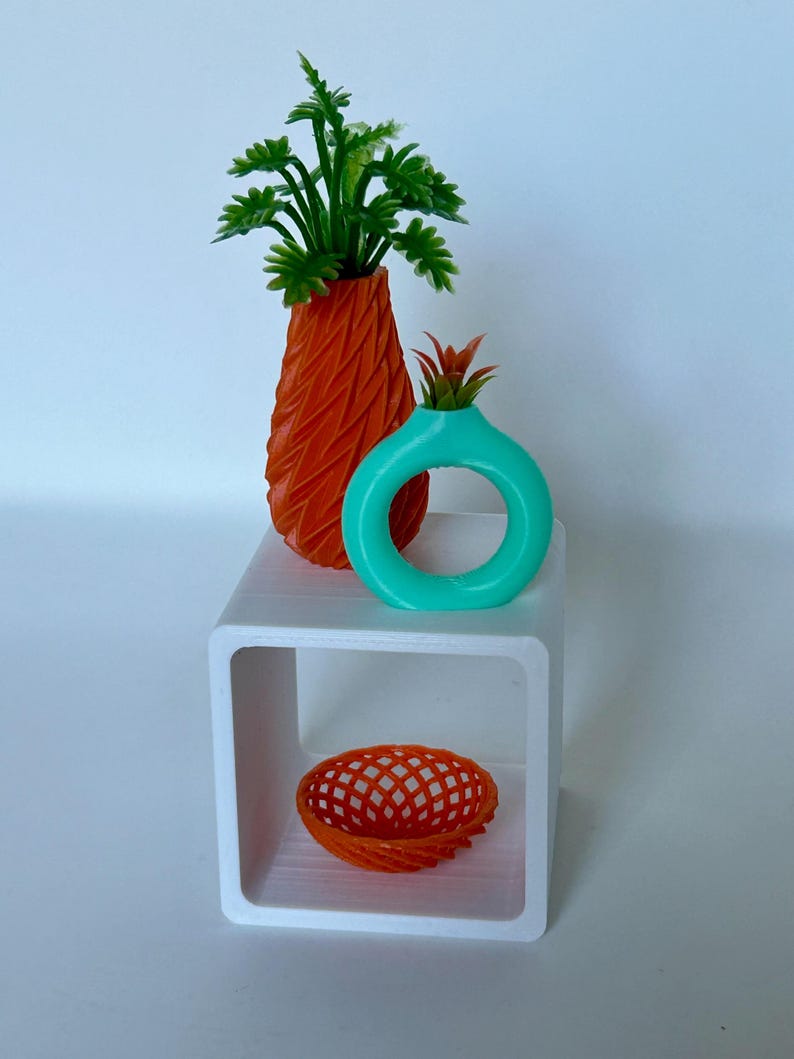 May include: A white cube shelf with three decorative items: a tall orange vase with a green plant, a turquoise vase with a red plant, and a small orange wire basket.