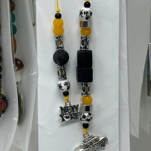 以下が含まれることがあります： 黄色と黒のビーズキーホルダー。シルバーのチャームには「NEW YORK」の文字が刻まれ、もう1つのシルバーチャームには黄色のタクシーが描かれています。